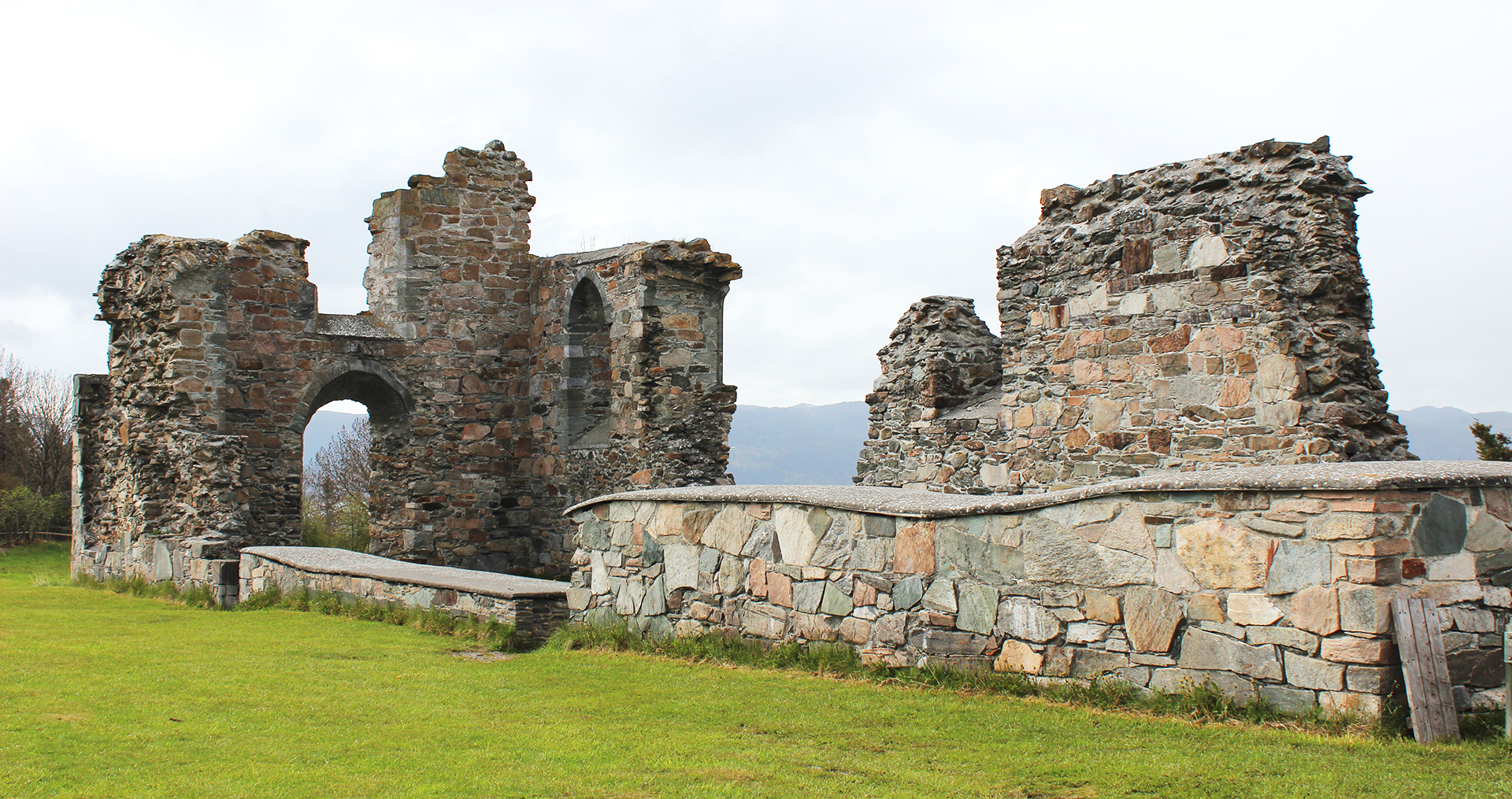 Ruinene av klosterkirken på Tautra, en steinbygning med høyde og detaljer av gammel arkitektur, omgitt av grønt gress og en bakketopp i bakgrunnen.