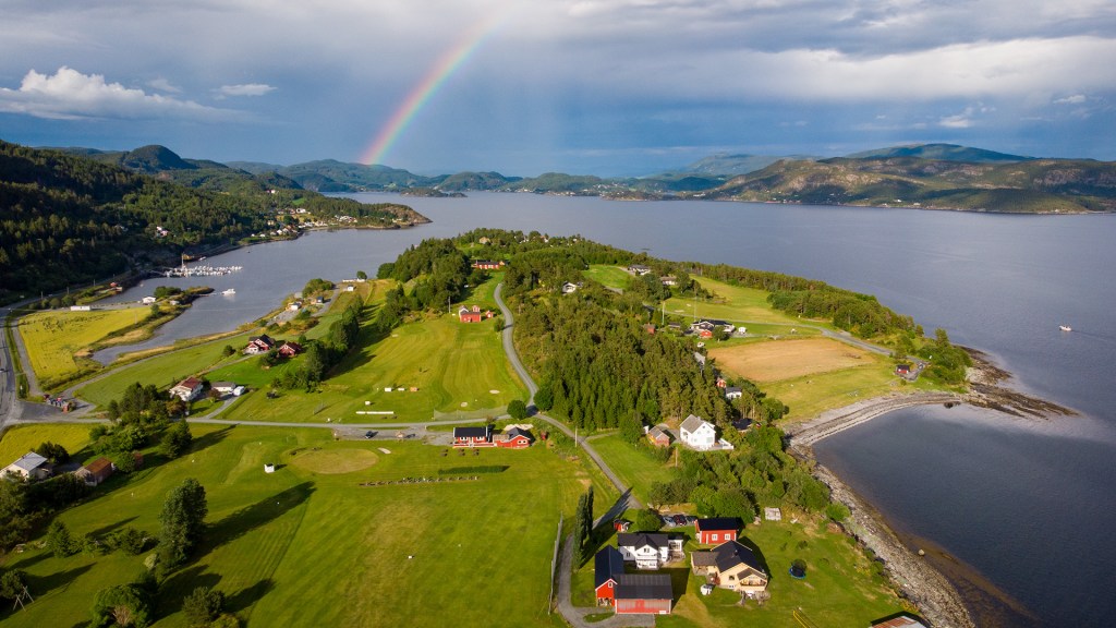 Luftfoto av frodig landskap på Frosta med grønne åser, hus og en lysende regnbue over havet.
