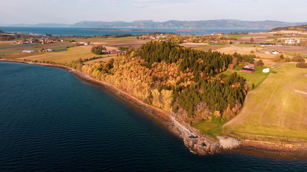 Autumn landscape at Frostating. A mix of forested hills and agricultural fields adjacent to the coastline, with distant mountains and a clear sky.