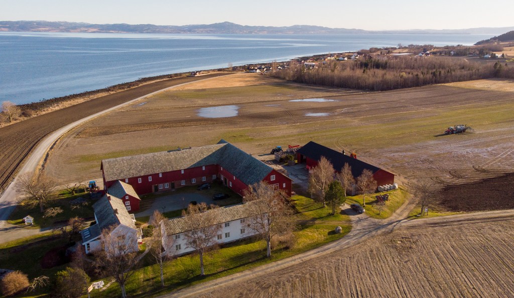 Panoramisk utsikt over en gård i Frostating utvalgte kulturlandskap, omgitt av åkre og fjorden i bakgrunnen. Det røde huset og bygningene danner en tradisjonell gård med karakteristiske tak og landskapsomgivelser.