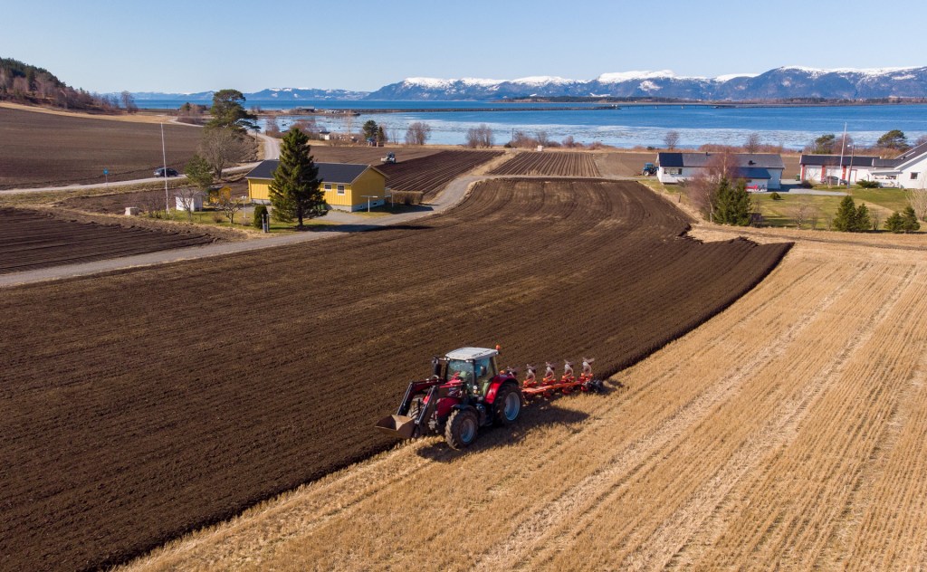 Luftbilde av en traktor som pløyer åkeren i Frosta med kystlinje i bakgrunnen og snødekte fjell.