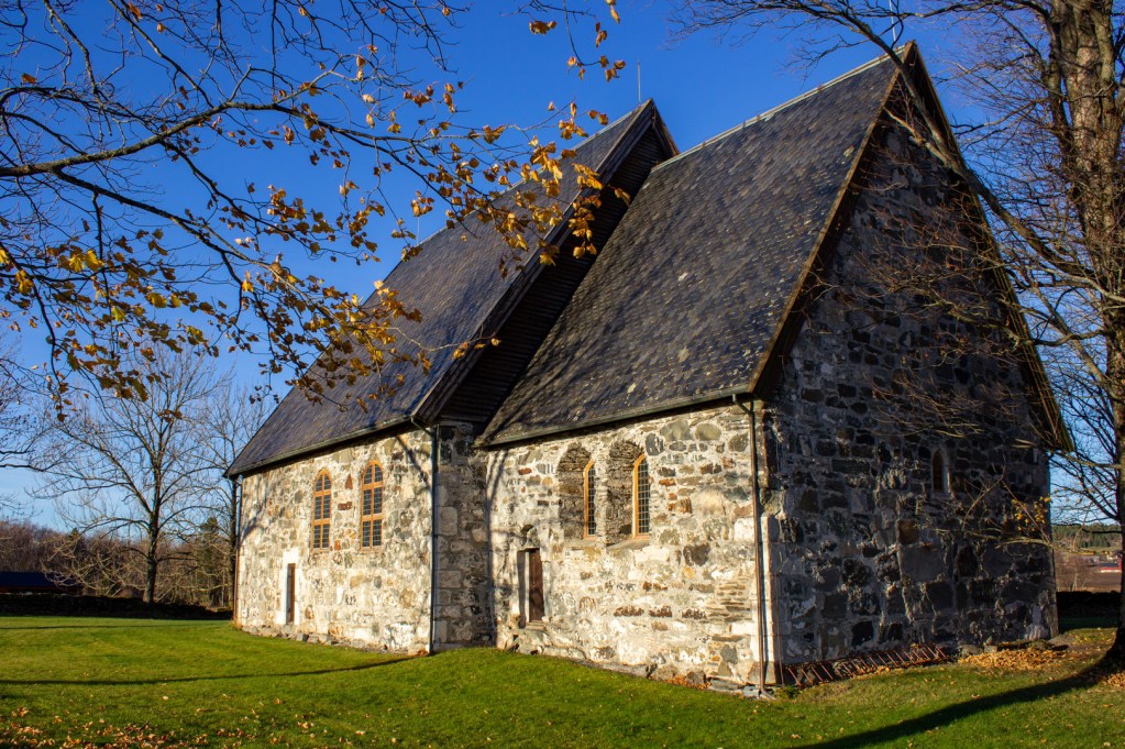 Logtun kirke, en steinkirke med gamle bygningselementer, omgitt av trær og gress, i et klart blått vær.