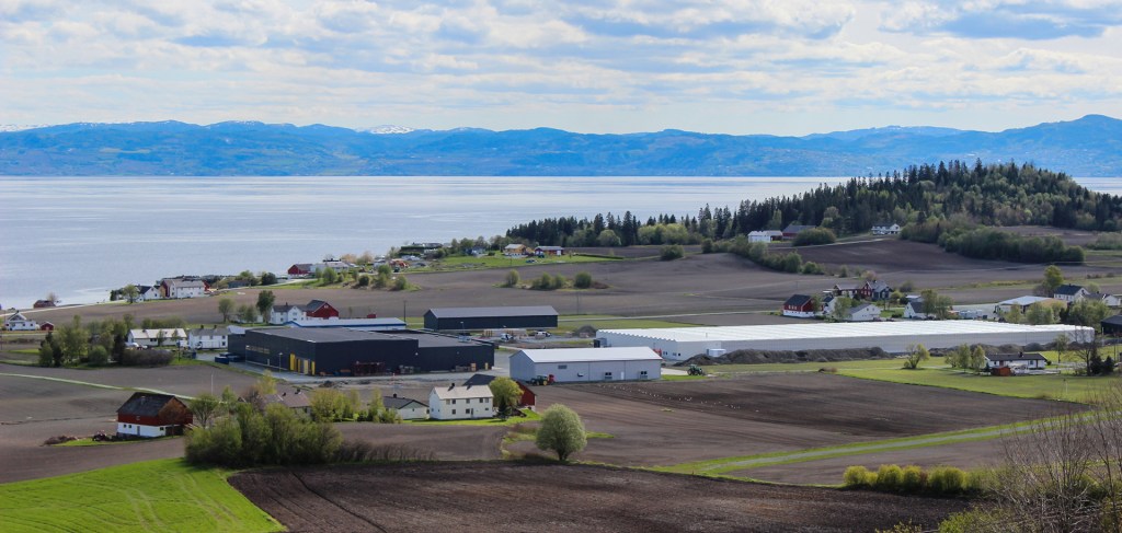 En panoramisk utsikt over landbrukslandskapet på Frosta, med bygninger og drivhus i forgrunnen, og innsjøen som strekker seg ut mot horisonten.