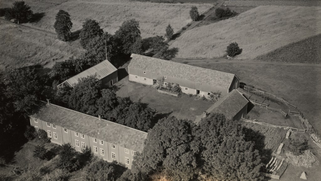 Luftbilde av en gård med firkanttun, omkranset av bygninger og vegetasjon, i et åpent landskap.