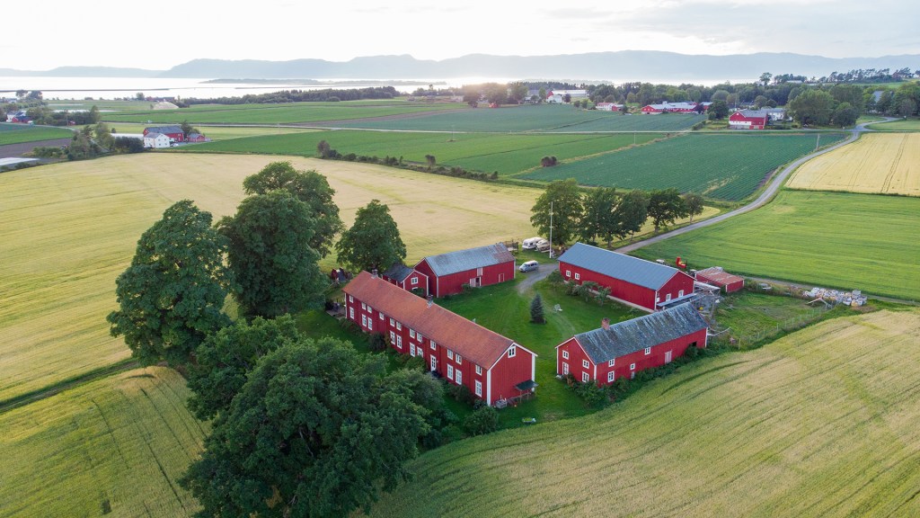 Luftbilde av en gård med rødmalte bygninger, omkranset av grønne åkre og trær, med fjord og fjell i bakgrunnen.