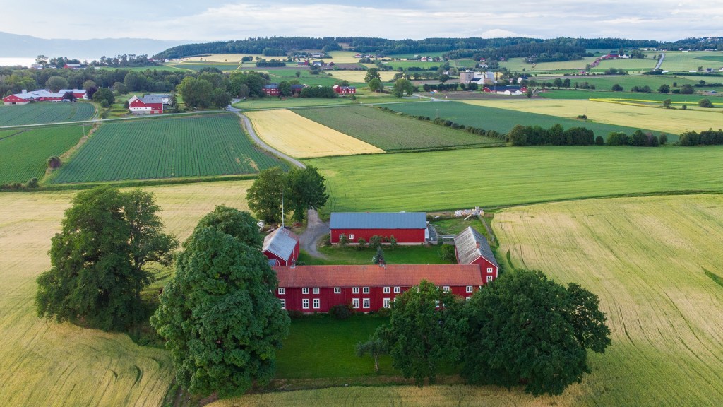 Oversiktsbilde av et gårdsanlegg med røde bygninger omgitt av grønne åser og jorder, med ulike avlinger i landskapet.