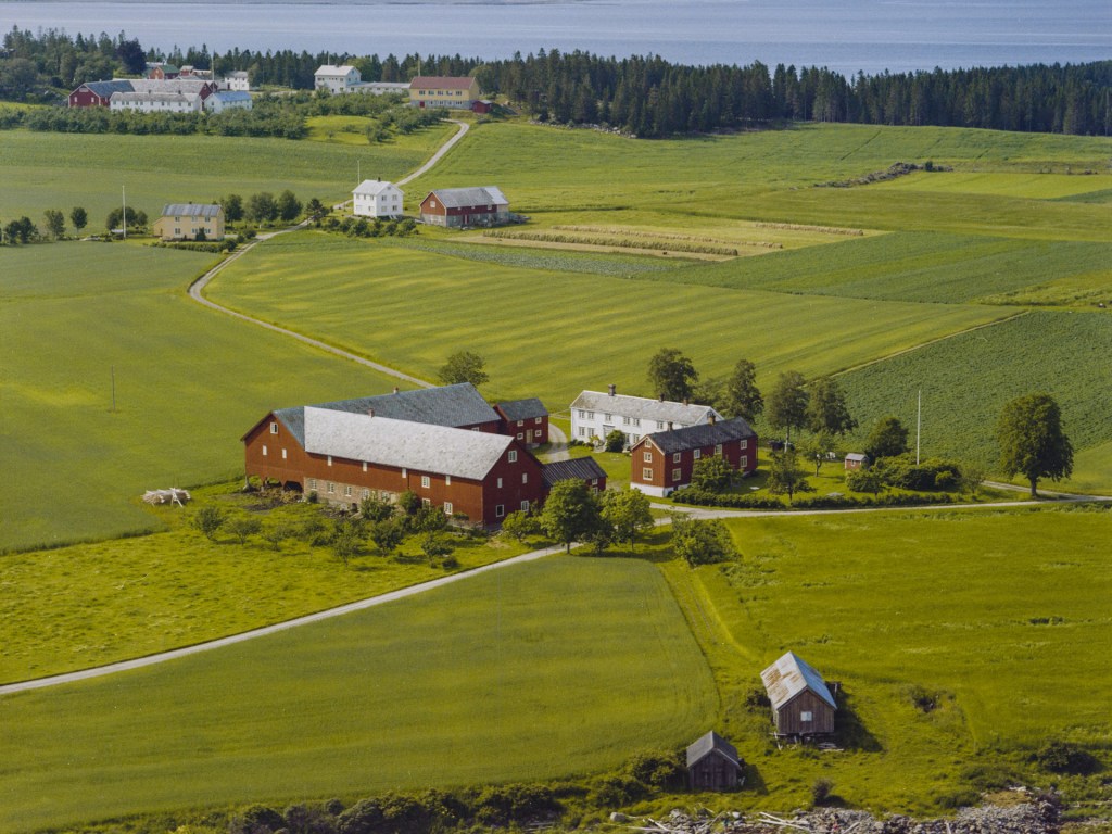 Luftbilde av gårdstun omgitt av grønne åkerområder og skogkledde bakgrunner, med flere bygninger i ulike farger synlige på landskapet.
