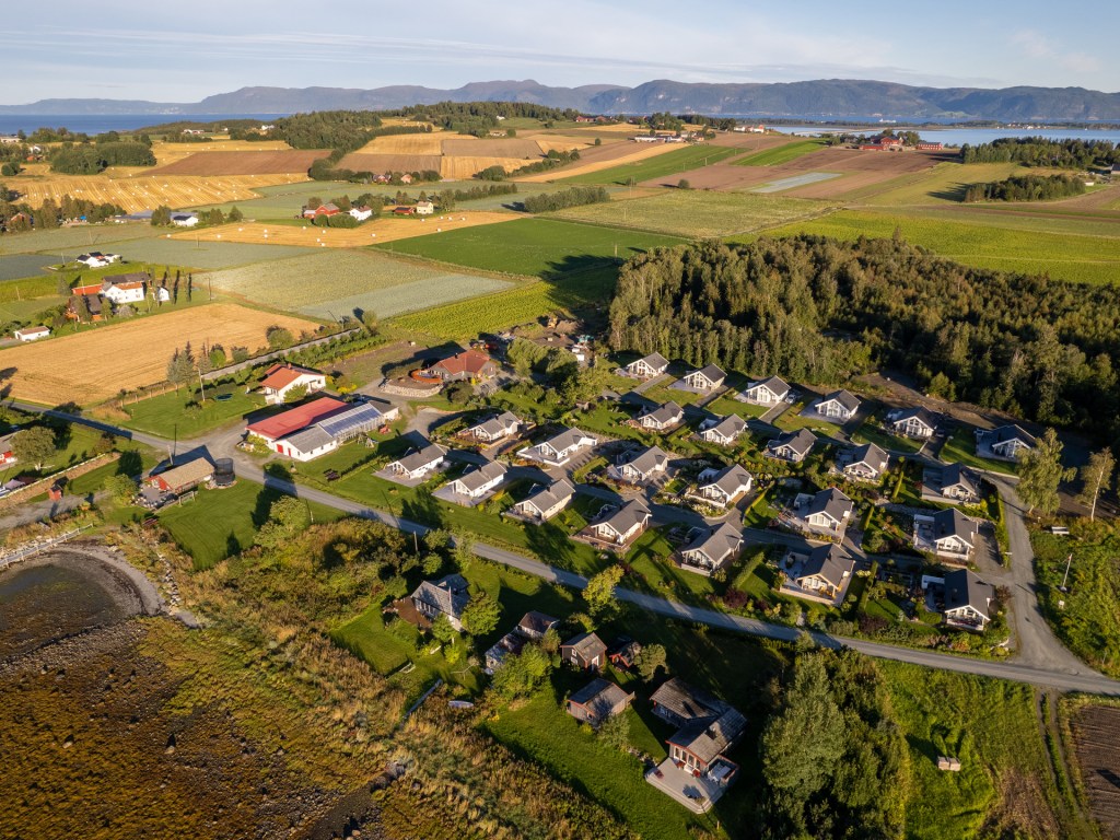 Luftbilde av landskapet i Frosta, Norge, som viser gårder, boliger og landbruksområder omgitt av grøntområde og fjorden i bakgrunnen.