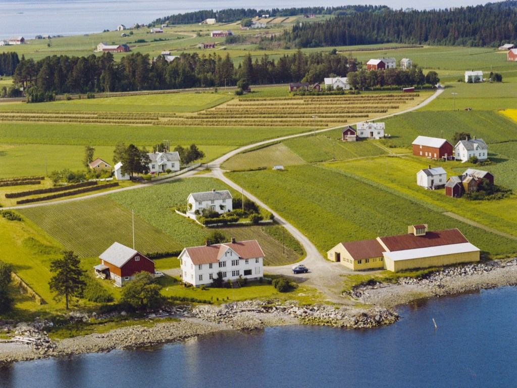 Luftbilde av et kulturlandskap med flere gårder, marker og trær, sett fra oven.
