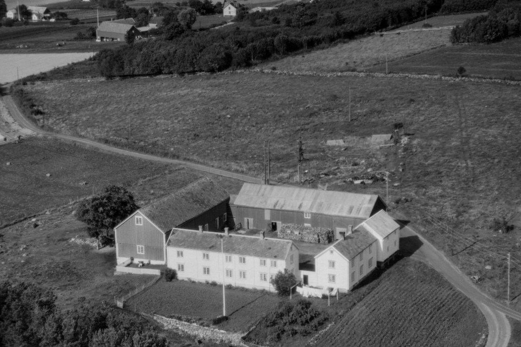 Luftbilde av en gård med flere bygninger, sett fra en høyde. Gården er omkranset av jordbrukslandskap, inkludert åkerland og trær, med en vei som går forbi.