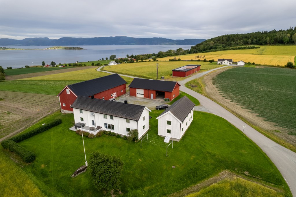 Landskapsbilde av en gård med moderne bygninger, beliggende nær vann med fjell i bakgrunnen, omkranset av grønne enger og åker.