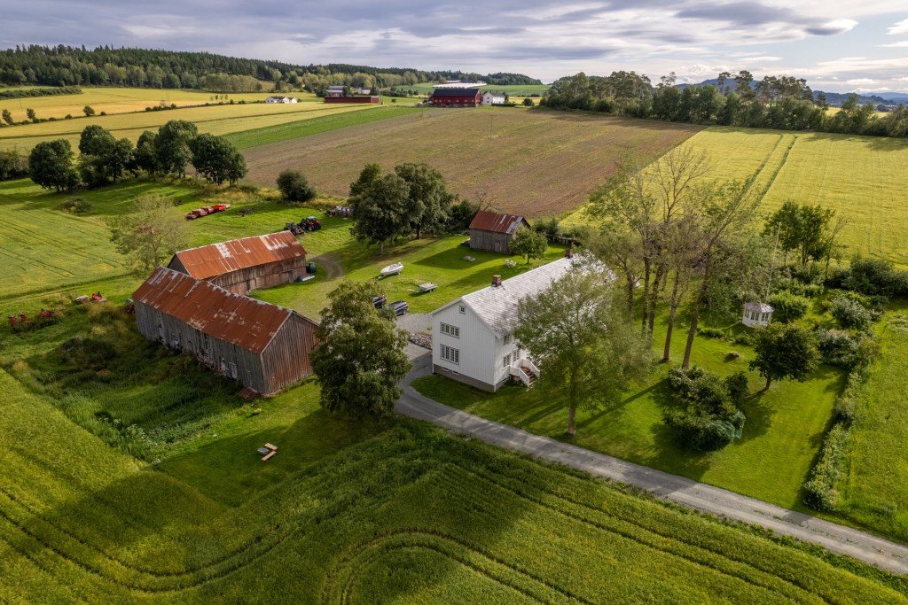 Luftbilde av en gård med flere bygninger, inkludert en hovedbygning og to eldre driftsbygninger, omringet av grønne åkre og trær.