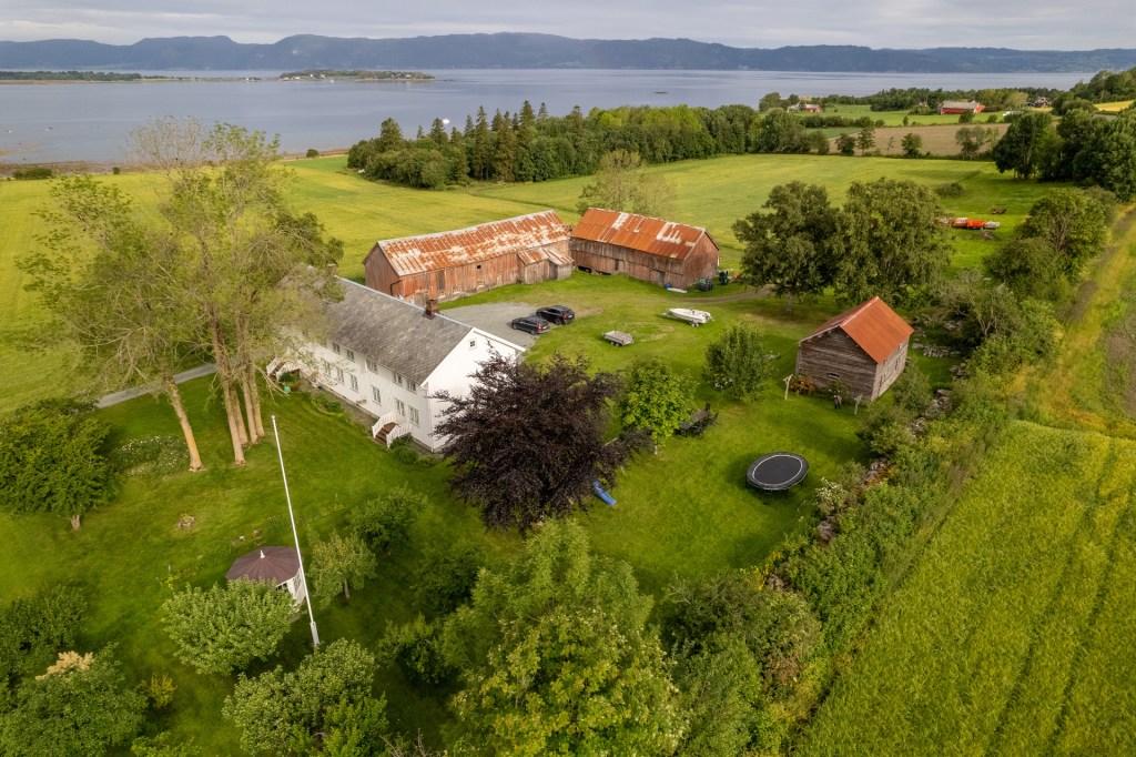 Luftbilde av gårdsanlegget med bygninger og grønt område, med en innsjø i bakgrunnen.