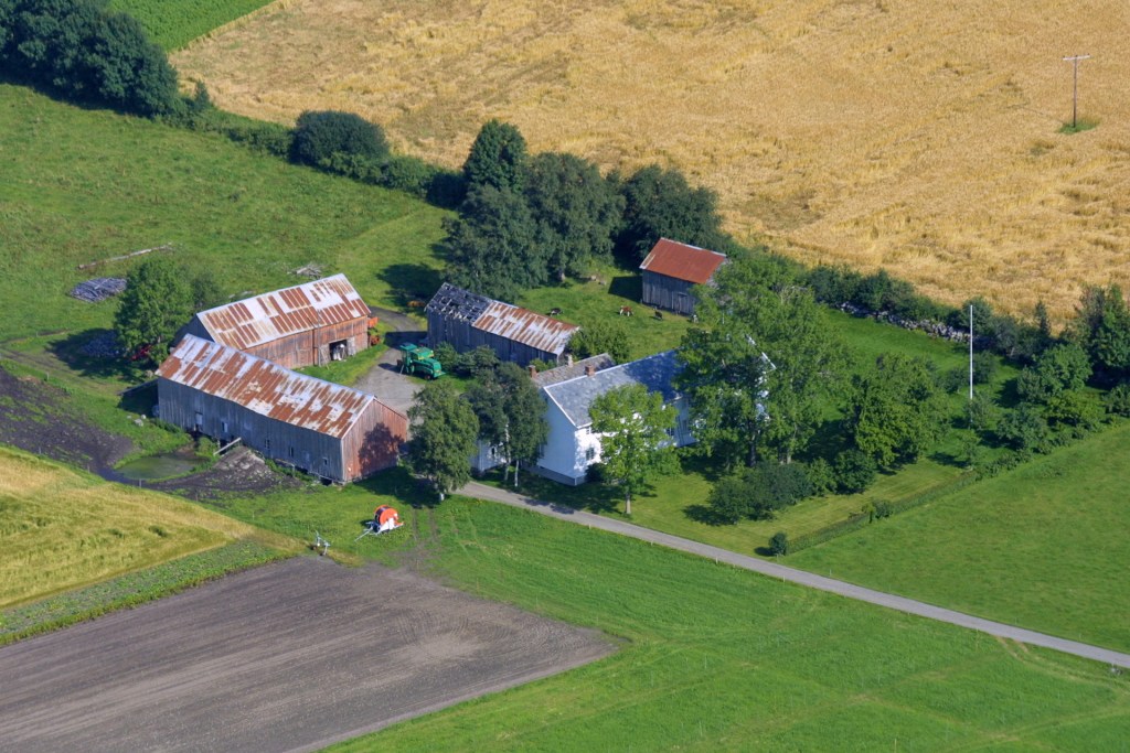Luftbilde av et gårdsanlegg med flere bygninger, inkludert stabbur og driftsbygninger, omgitt av grønne marker og åkerlandskap i en landlig setting.