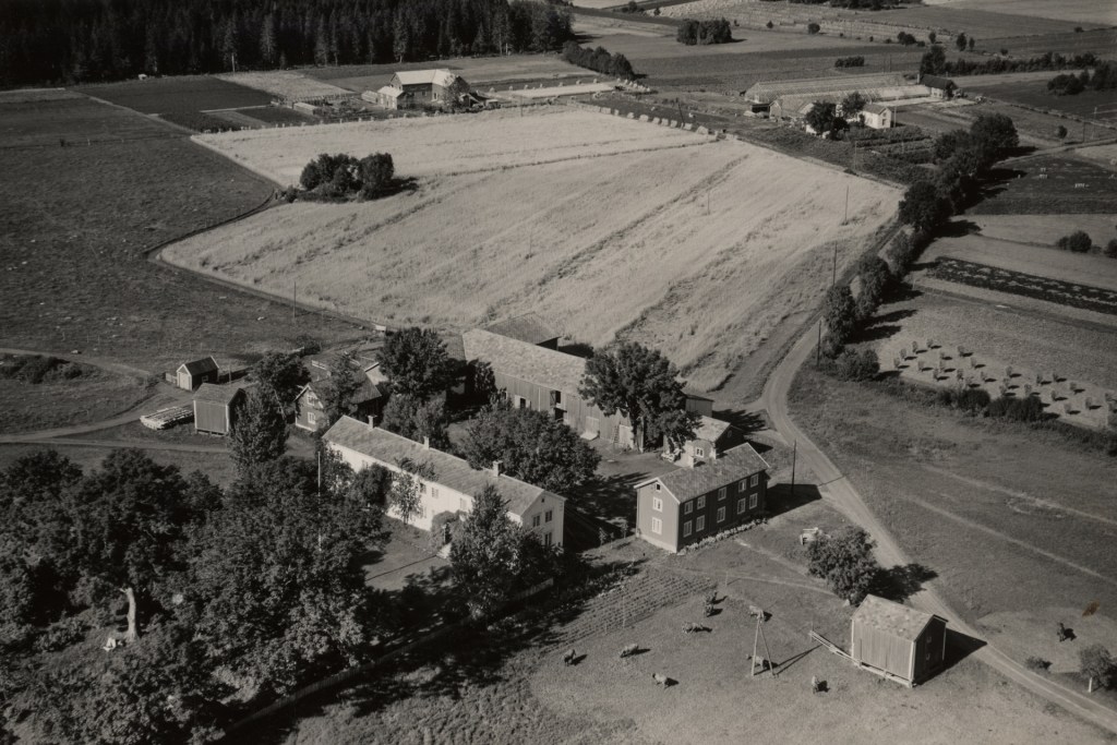 Luftbilde av et tradisjonelt gårdstun med firkanttun, omgitt av naturlandskap og dyrkede marker i bakgrunnen.