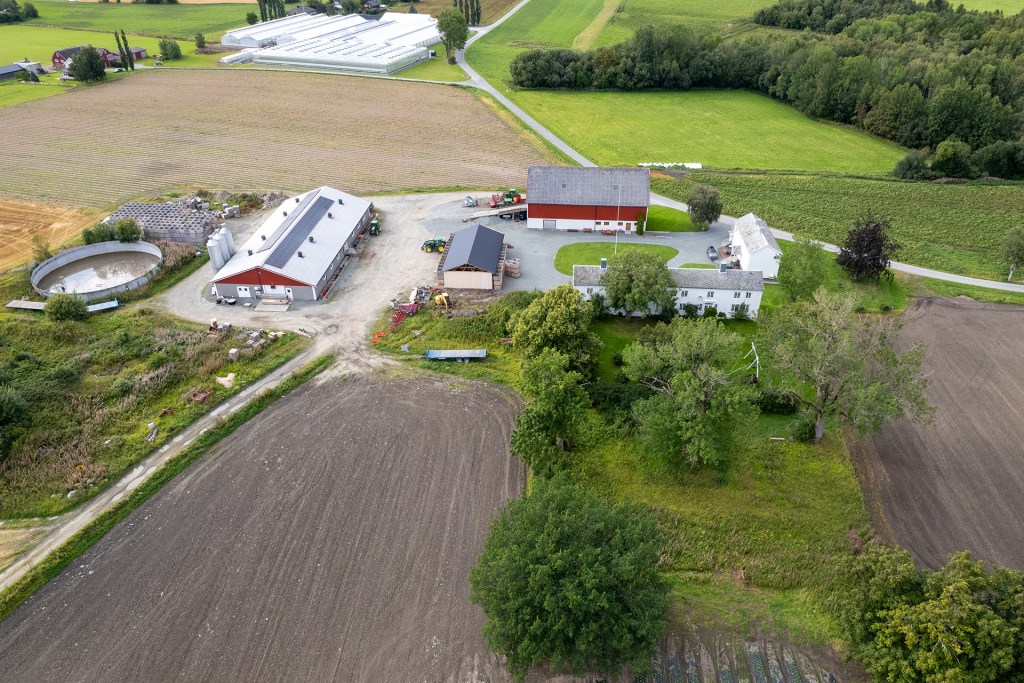 Luftbilde av en gård med moderne driftsbygninger og veksthus, omgitt av jorder og grønt landskap.