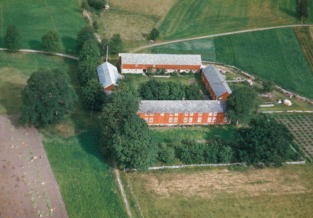 Oversiktsbilde av et gårdstun med røde bygninger omgitt av grønt landskap og mark.