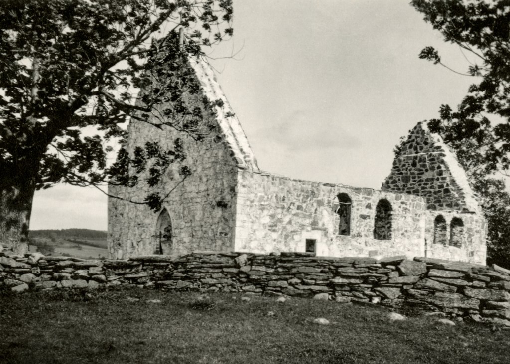 En svart-hvitt fotografering av ruiner av en steinkirke med utsikt over landskapet og trær i forgrunnen.