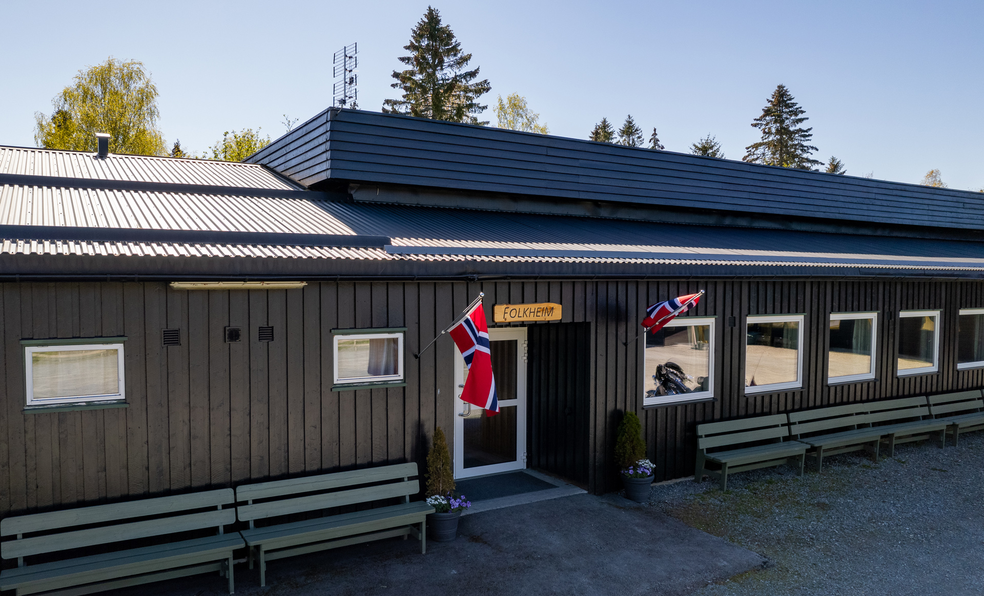 Fasaden til en bygning med skråtak, merket 'Folkheim', med det norske flagget heist på to flaggstenger, omgitt av grønne benker og blomster i potter.