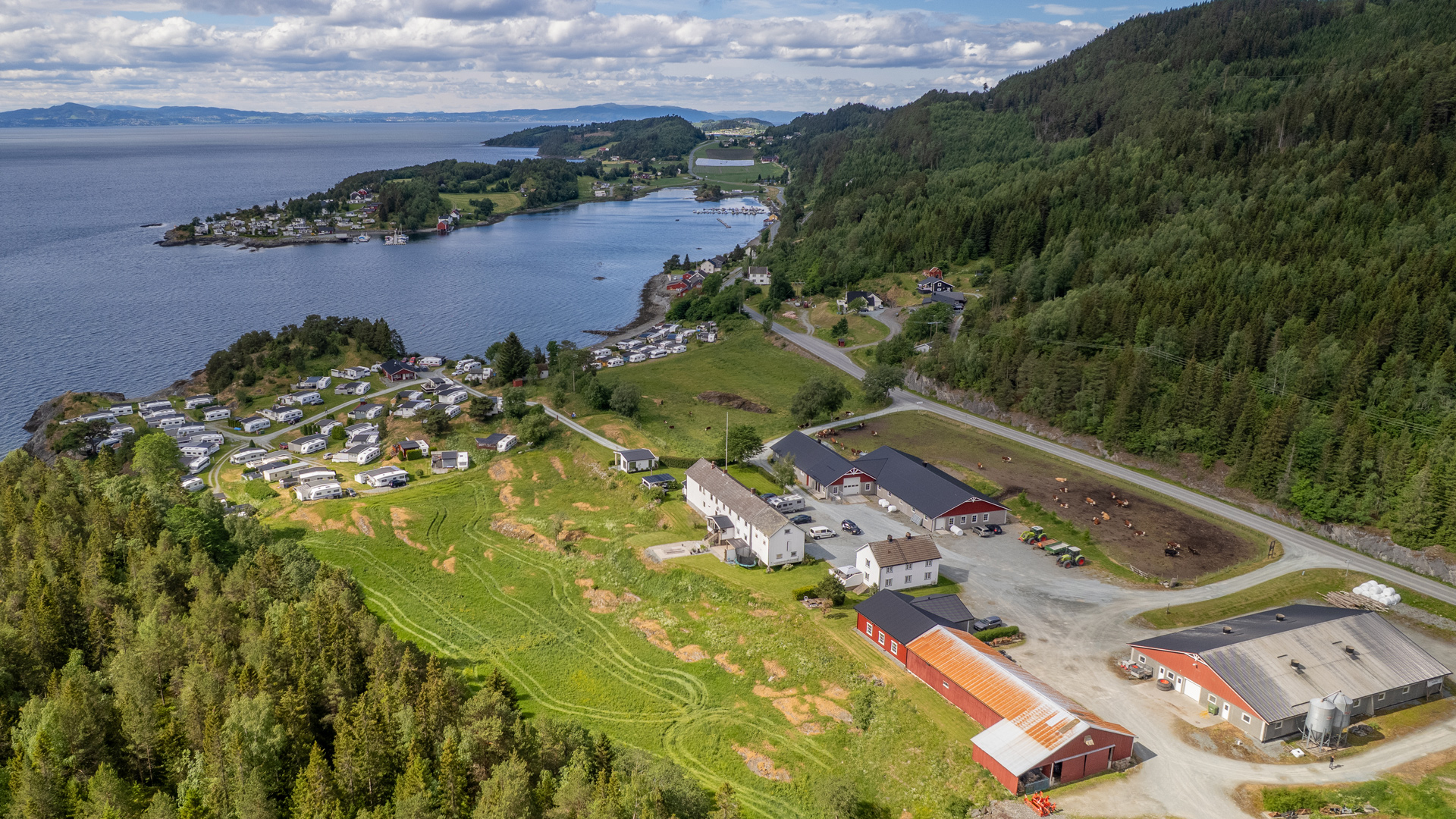 Luftbilde av et campingområde ved sjøen, med campingvogner og hytter omgitt av frodig natur og fjordlandskap.
