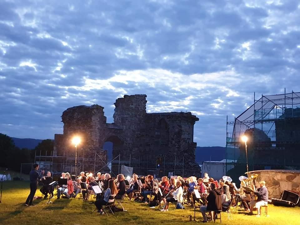 En konsert med et stort orkester som spiller foran en gammel ruin under en kveldshimmel med skyer.
