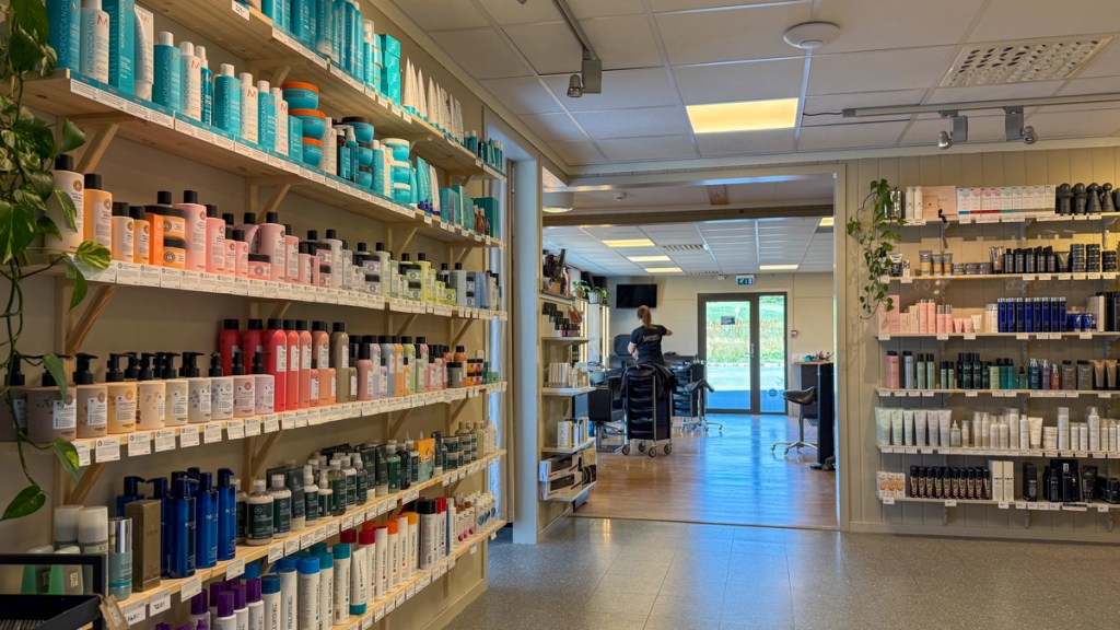 Interiørbilde av en frisørsalong med hyller som inneholder forskjellige hårprodukter, og en frisør som jobber med en kunde i bakgrunnen.