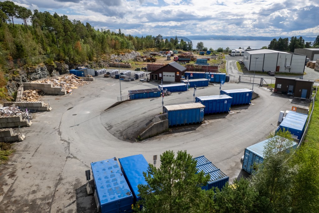 Oversiktsbilde av et gjenvinningsanlegg med flere blå containere, et verksted og skog i bakgrunnen.