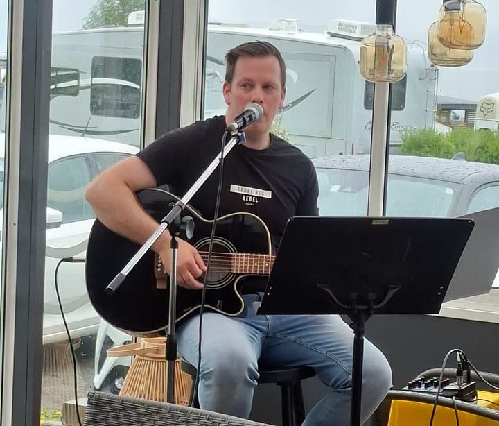 Mann som spiller akustisk gitar og synger på en scene, med mikrofon foran seg. Det er et bord og stoler i forgrunnen, og utsikt mot biler og campingvogner utenfor vinduet.